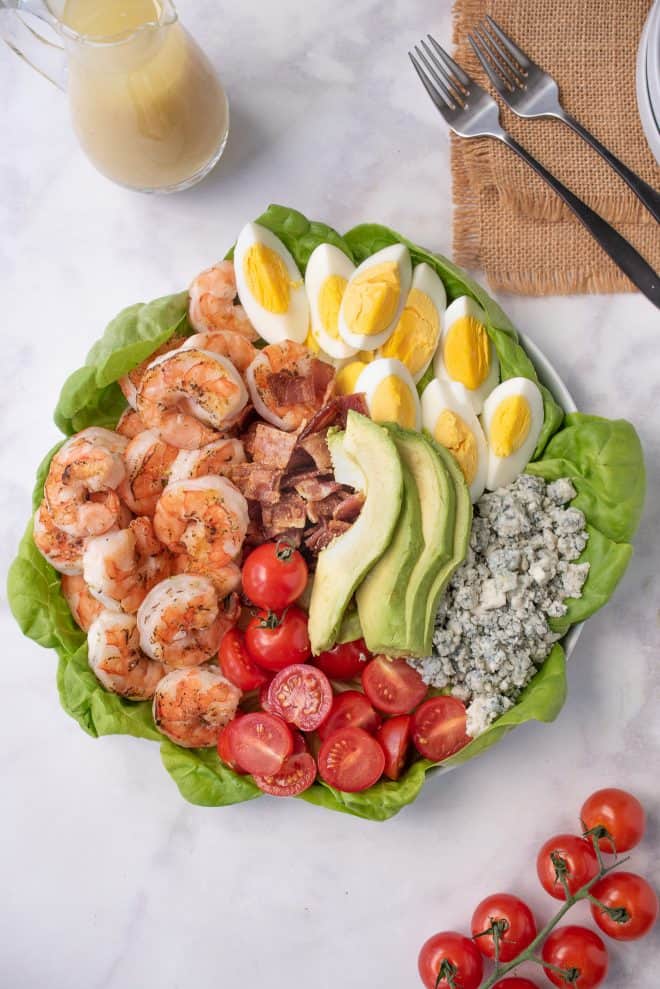 A large platter with lettuce leaves, grilled shrimp, tomato, bacon, egg, avocado and blue cheese