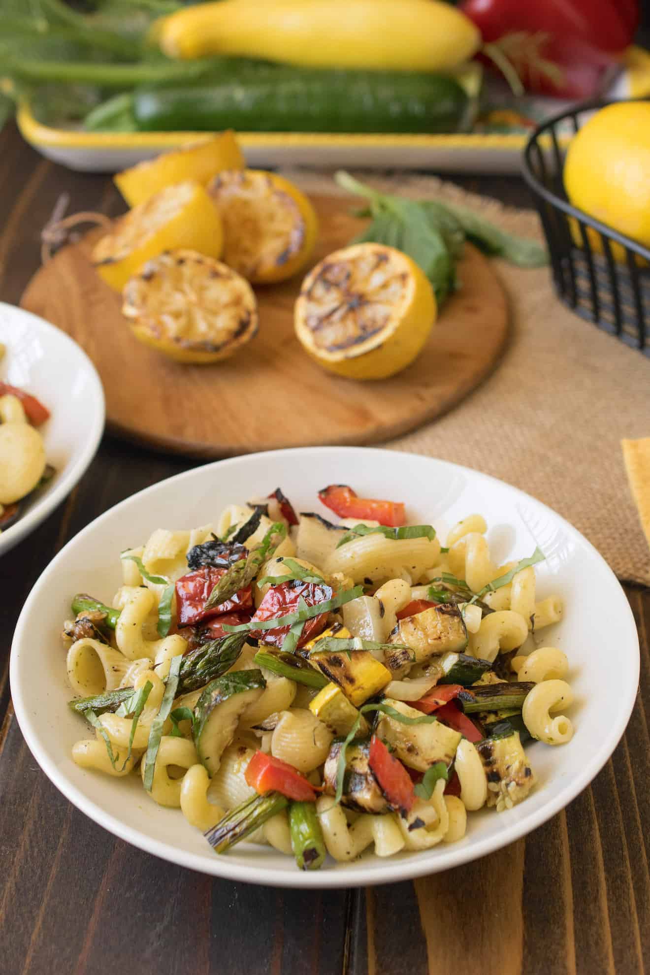 Grilled spring vegetables mixed with pasta and lemon dressing in a white bowl with grilled lemons in the background