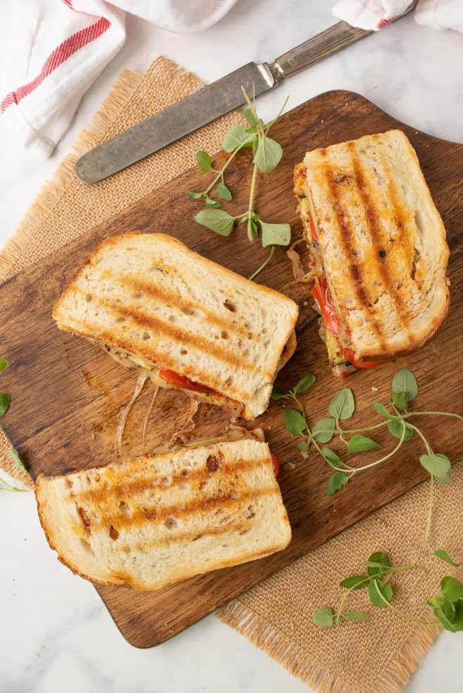 3 halves of a Grilled summer vegetable panini from above on a board with a knife and fresh herbs