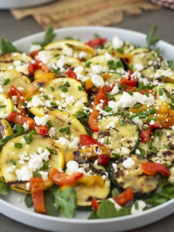 Zucchini and yellow squash on a white round platter with arugula peppers and feta