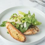 A Caesar salad on a white plate with a chicken breast and bread crostini