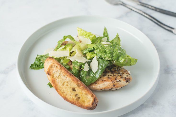 A Caesar salad on a white plate with a chicken breast and bread crostini