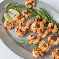 Skewers of grilled shrimp on a grey oval plate with fresh lime wedges and rosemary