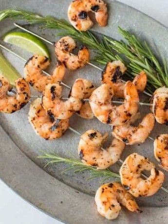 Skewers of grilled shrimp on a grey oval plate with fresh lime wedges and rosemary
