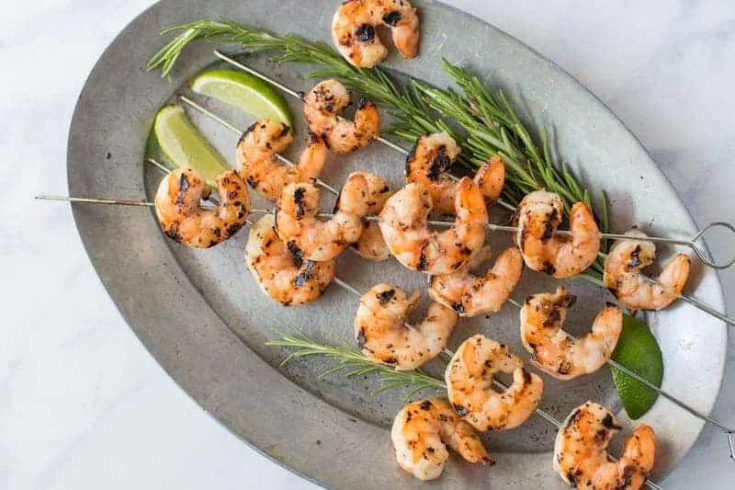 Skewers of grilled shrimp on a grey oval plate with fresh lime wedges and rosemary