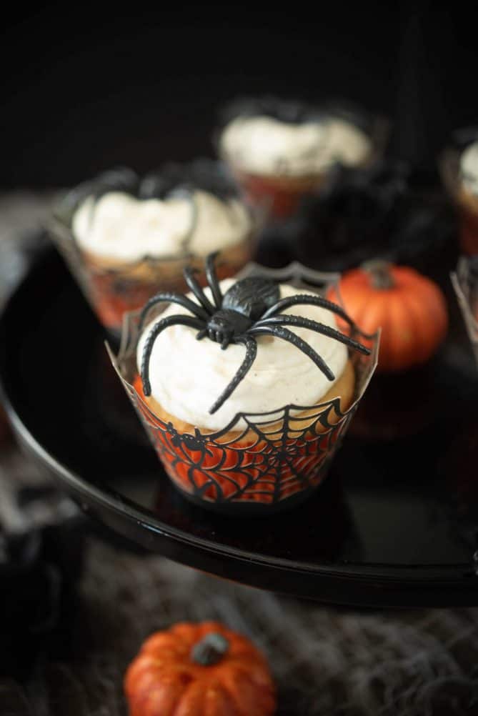 A closeup of a cupcake in a spider web muffin cup topped with a large plastic spider with mini orange pumpkins around