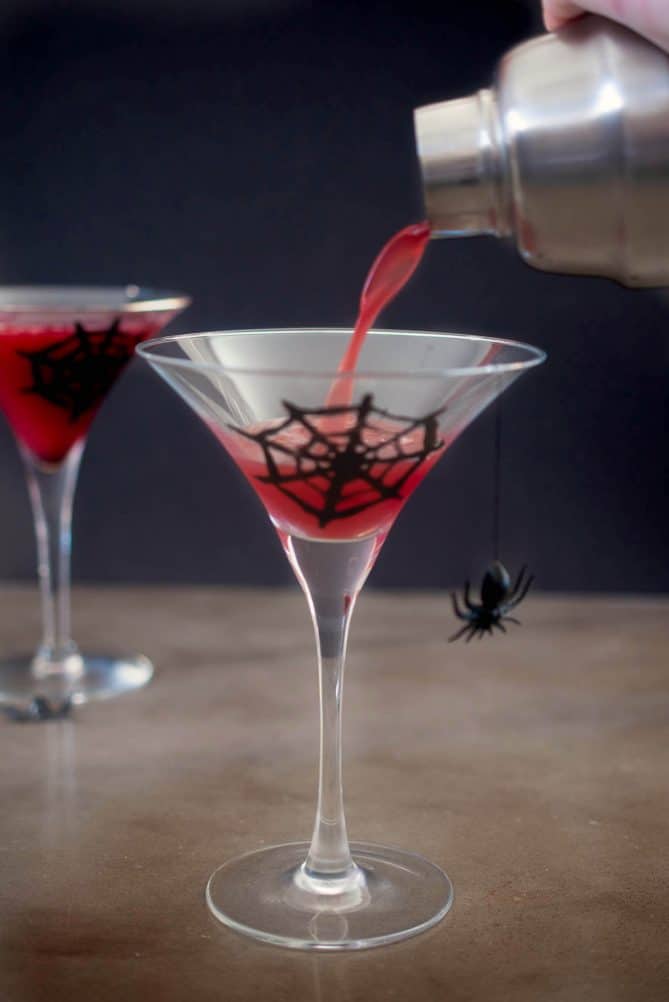 Pouring red juice into a martini glass that has a web painted on it and a plastic spider hanging from the rim