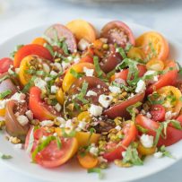 Multi-color sliced tomatoes on a round white plate topped with toasted pistachios, feta and fresh basil