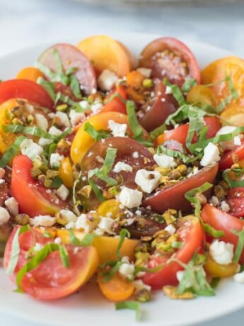 Multi-color sliced tomatoes on a round white plate topped with toasted pistachios, feta and fresh basil