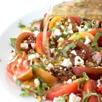 Pouring dressing onto a platter of sliced tomatoes with pistachios, feta and fresh basil