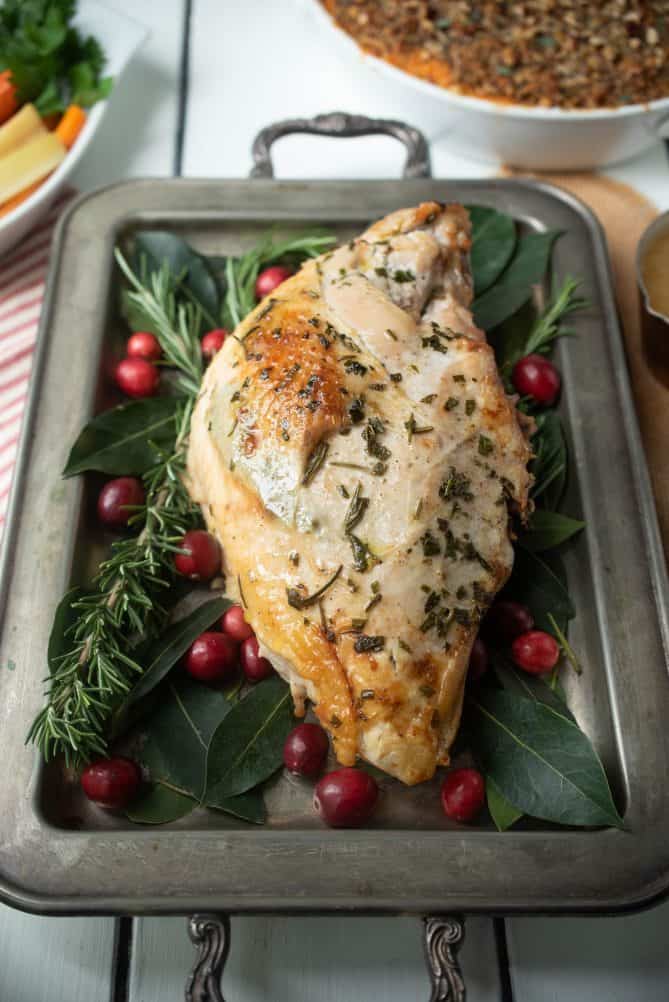 A perfectly browned roasted turkey breast covered with herbs on a serving tray with herbs and cranberries with sweet potato casserole in the background