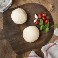 2 pizza dough balls on a board with tomatoes and basil