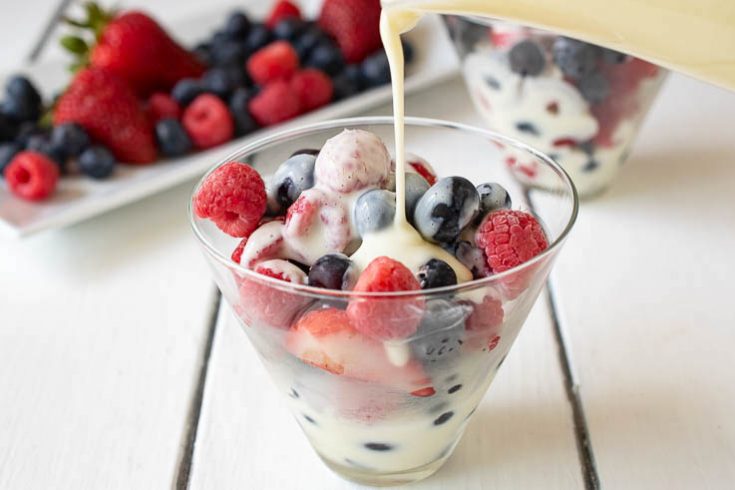 Frozen raspberries, blueberries and strawberries in a glass bowl with custard being poured over
