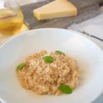 Simple risotto piled up in the center of a white bowl with a glass of white wine and a wedge of Parmesan cheese
