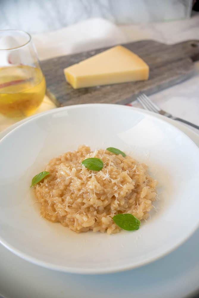 Simple risotto piled up in the center of a white bowl with a glass of white wine and a wedge of Parmesan cheese