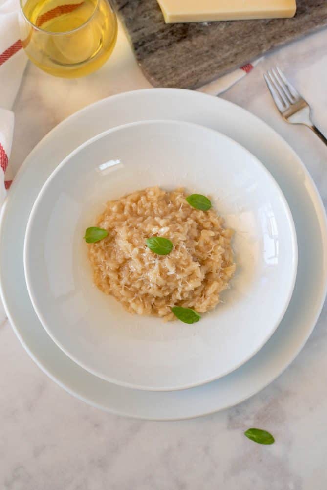 A large white plate with a white pasta bowl filled with risotto garnished with small basil leaves