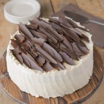 A cream colored frosted cake adorned with chocolate shards on a cake stand