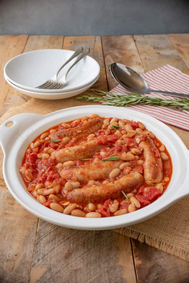 A round white serving bowl with sausage and beans with 2 bowls and forks