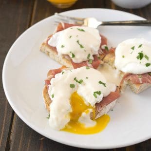 A poached egg on top of prosciutto and ciabatta bread cut open with yellow yolk running out