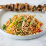 Noodle salad with vegetables on a white plate with grilled chicken skewer