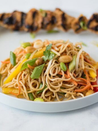 Noodle salad with vegetables on a white plate with grilled chicken skewer