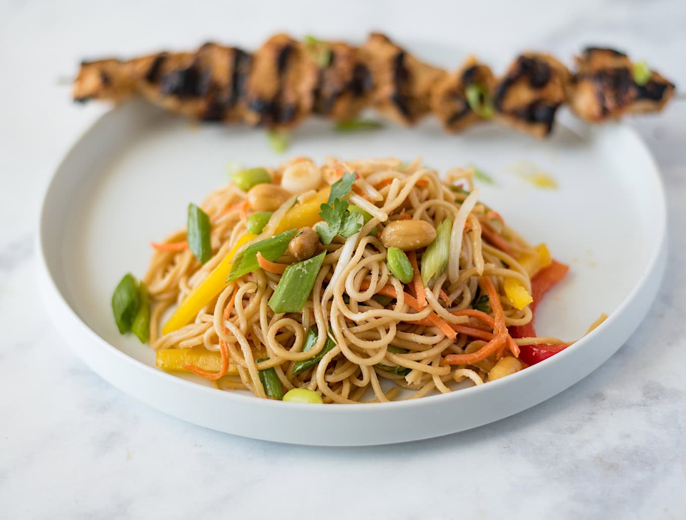 Vegetable noodle salad on a white plate with grilled chicken skewer