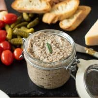 A jar of mushroom pâté on a slate board with cherry tomatoes, cheese and bread