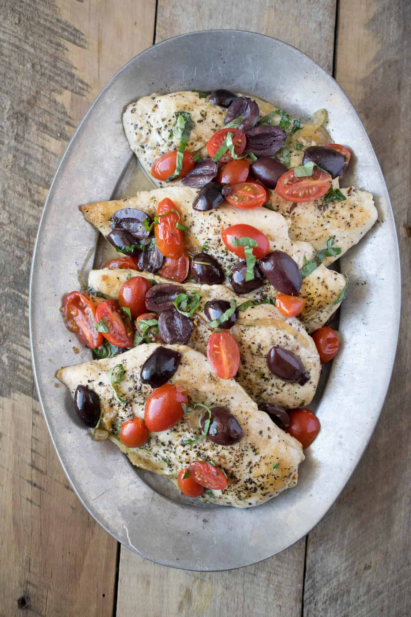 An oval serving dish with chicken breasts topped with herbs, Kalamata olives and tomatoes