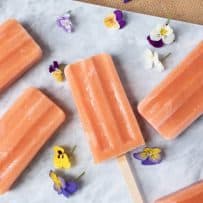 5 peach popsicles laying down on a marble board surrounded by edible purple, yellow and white pansy flowers
