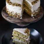 A whole pistachio cake on a wood cakes stand with a slice next to it on a black plate