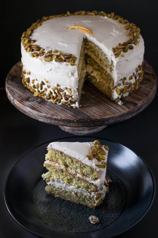 A whole pistachio cake on a wood cakes stand with a slice next to it on a black plate
