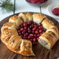 A puff pastry sausage roll wreath on a round board with 1 missing with fresh cranberries and a cranberry apple sauce in the background