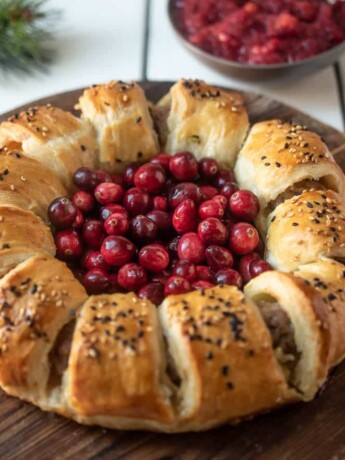Puff pastry rolled into a wreath shape filled with pork sausage on a board with fresh cranberries in the center