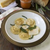 A white bowl of pumpkin ricotta ravioli garnished with sage and cheese and a side of marinara sauce