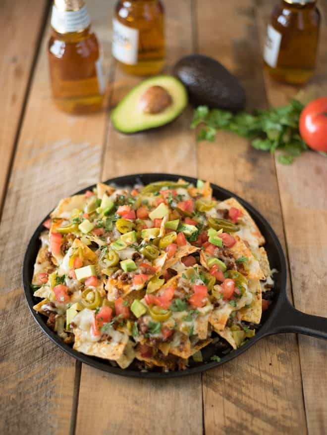 Nachos topped with tomato, jalapeño, avocado and cilantro with bottles of beer in background