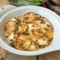 A white round dish of chicken piccata with lemon slices and capers.