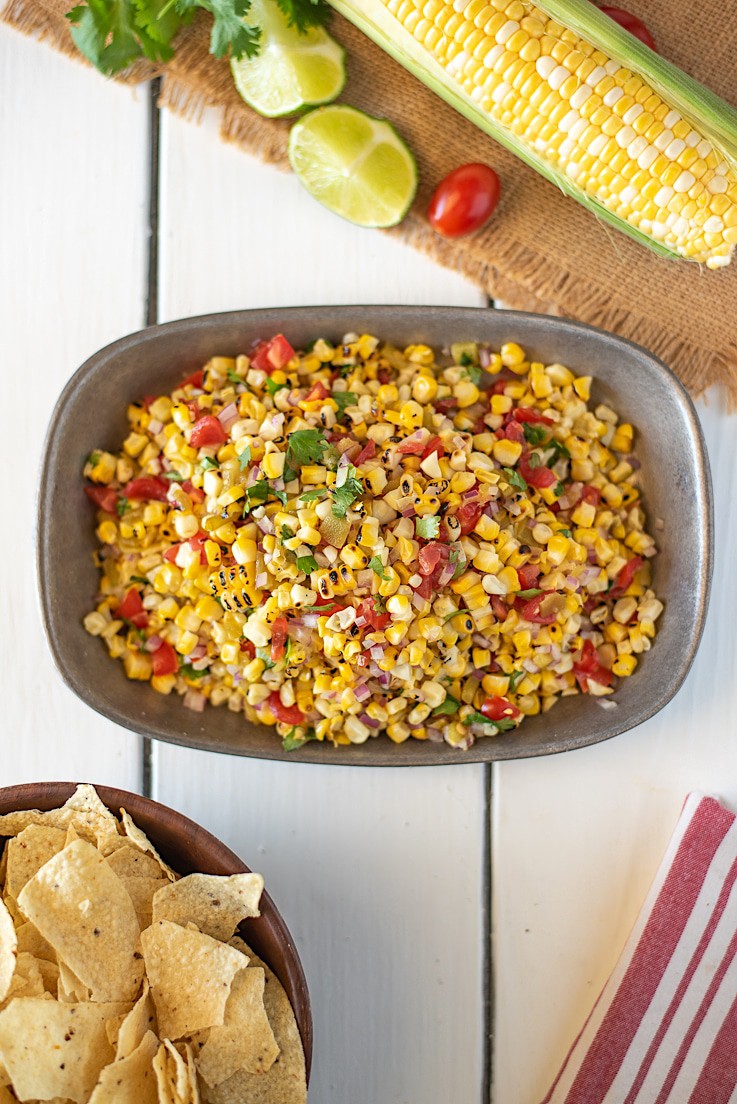 An overhead image showing the colorful salsa in a pewter bowl with tortilla chips, lime wedges and corn on the cob