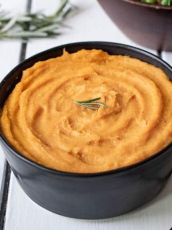 Vibrant orange puree in a black bowl garnished with fresh rosemary with a serving spoon