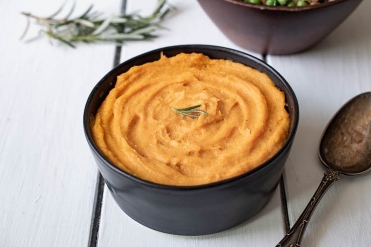 Vibrant orange puree in a black bowl garnished with fresh rosemary with a serving spoon