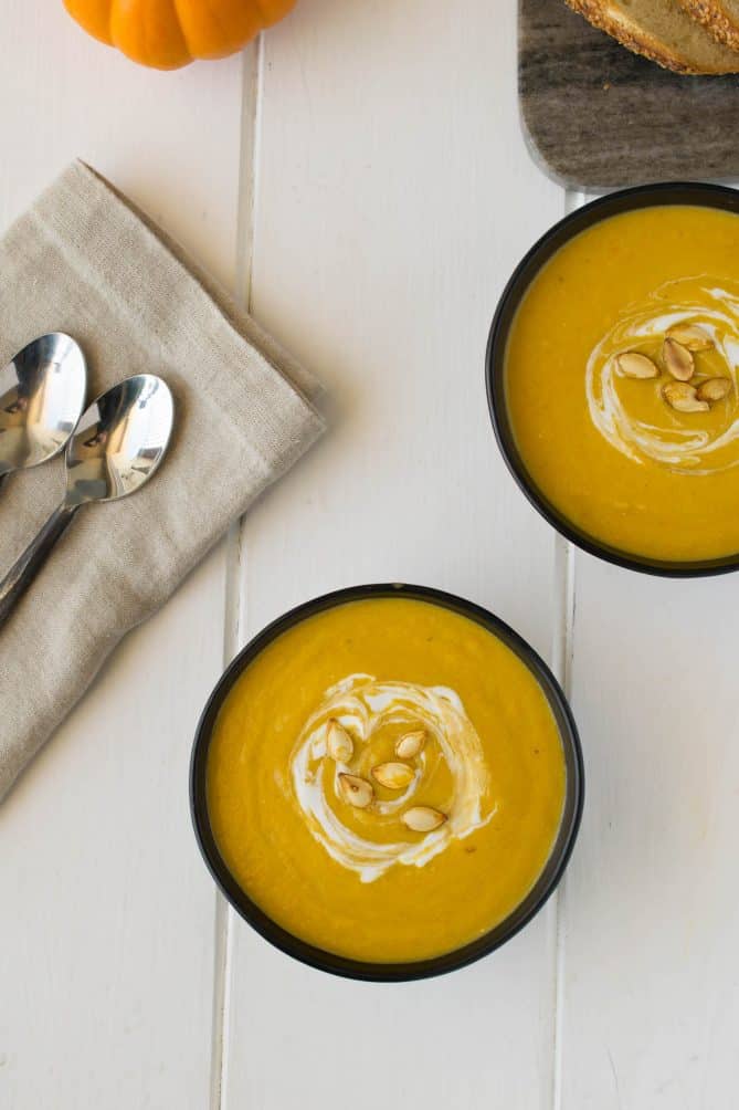 2 bowls of pumpkin and root vegetable soup viewed from overhead with 2 spoons and a linen napkin