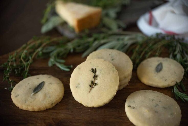Herb and parmesan shortbread crackers displayed on a board with fresh herbs