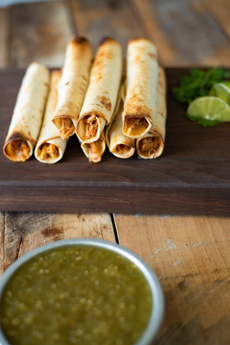 Shredded chicken baked taquitos stacked on a serving board with fresh lime and salsa verde