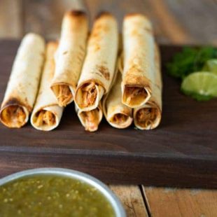 Taquitos on a board with lime wedges and green salsa