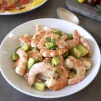 A white bowl filled with cooked shrimp, avocado and salad dressing with a large serving spoon