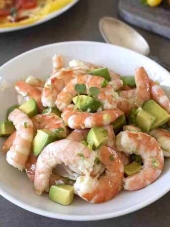 A white bowl filled with cooked shrimp, avocado and salad dressing with a large serving spoon