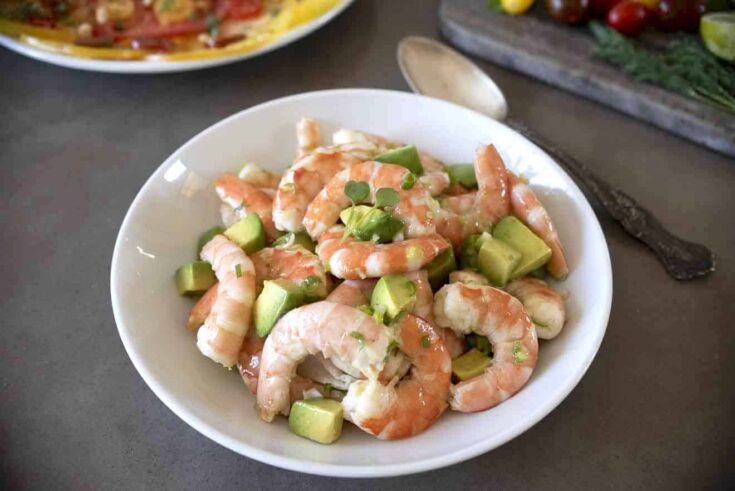 A white bowl filled with cooked shrimp, avocado and salad dressing with a large serving spoon