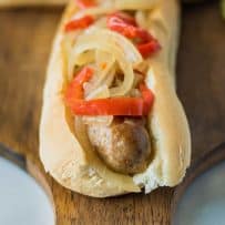 A closeup of a slow cooker beer bratwurst on a bun topped with onions and peppers