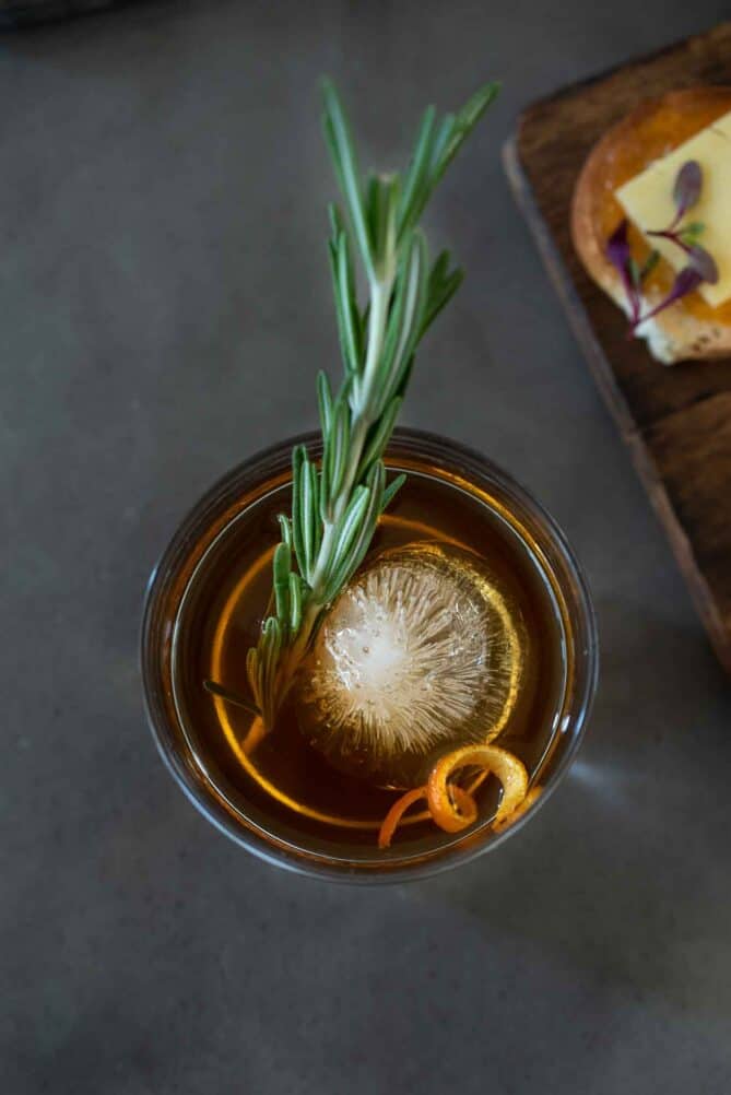 A Manhattan cocktail from overhead with a sprig of rosemary and curled orange peel