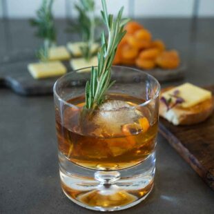 A smoked Christmas Manhattan drink with a large ice cube, orange peel and rosemary sprig