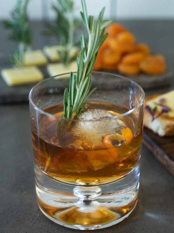 A smoked Christmas Manhattan drink with a large ice cube, orange peel and rosemary sprig